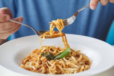 Eller, yeşil yapraklarla süslenmiş yuvarlak bir tabağa çatal ve kaşıkla spagetti koyar.