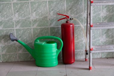 Duvarın önünde bir envanter seti var. Plastik yeşil sulama kutusu ve fayanslı kırmızı yangın söndürücü.