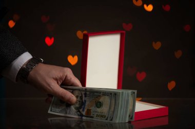 Sevgililer günü hediyesi. Bir el kırmızı bir kutuya bir tomar dolar koyar. Sarı kalpler arkada parıldıyor.