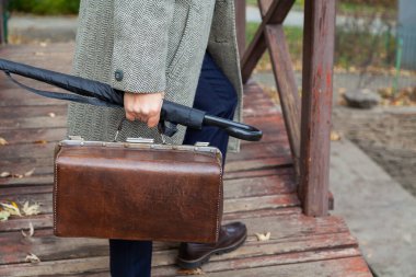 Köprüdeki beyefendi. Şık ayakkabılı ve gri paltolu bir adam sonbahar parkında ahşap bir köprü boyunca yürüyor. Elinde bir şemsiye ve valiz.