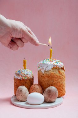Pembe arka planda iki Paskalya pastası ve yumurta standı. Mumlar Paskalya pastalarının içine konur. Bir el kibritle bir mum yakar. Ön planda beyaz çiçekli bir ağaç dalı var.
