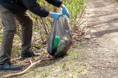 Doğayı çöpten kurtarıyoruz. Mavi lastik eldivenli ekolojist ön taraftaki siyah plastik torbada çöp topluyor.