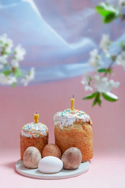 イースターはまだ生きてる ピンクの背景に2つのイースターケーキと卵が丸いスタンドに立っています キャンドルはイースターケーキに挿入されます 青い布のシンボルの空と白い花の枝を上から見る — ストック写真