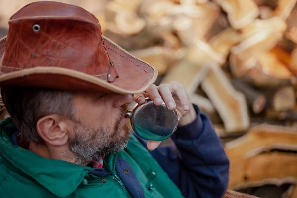Profile of a man in a leather hat who drinks from a round metal flask against a background of chopped firewoo