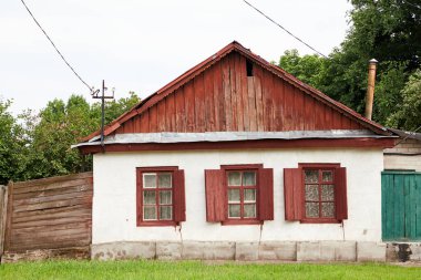 Bir katlı, ahşap çatılı ve yaz günü tavan arası olan bir ev. Beyazlatılmış duvarda açık ahşap panjurlu üç pencere var.