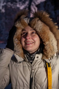 Kapüşonlu açık renk ceketli genç bir kadın kış caddesinde cep telefonuna bakıyor. Doğal kusurlarıyla rötuşsuz bir kadın portresi.