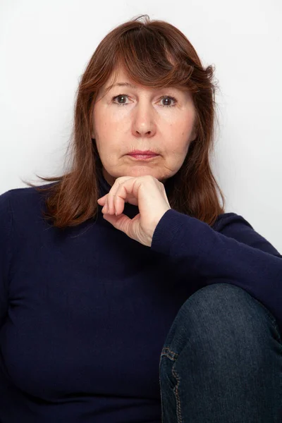 Portrait of a woman without retouching sitting on a light solid background. An elderly beautiful woman in a dark sweater looks into the fram