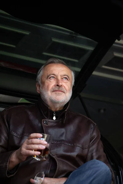 After the trip. An elderly man with a gray beard in a leather jacket holds in his hand a glass of whiskey against the background of an open trunk of a car in a garag