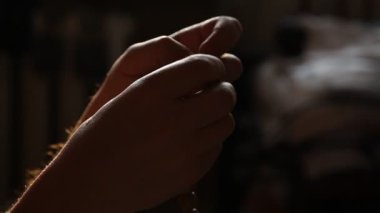 Siyah arka planda, tahta bir tespihi iki elle sıralayan bir adam. Yukarıdan loş bir ışık düşüyor