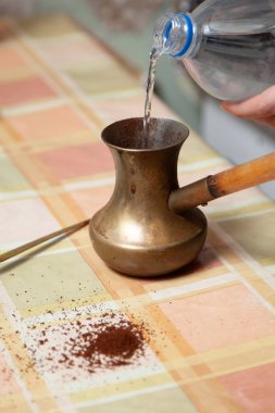 Doğal kahve yaparız. Eller plastik bir şişeden pirinç bir kaba su döker. Kareli masanın üzerinde uzun bir kaşık var.