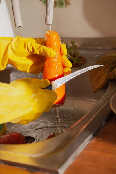 Hands in yellow rubber gloves cut the carrot with a thin knife in the weight. Close-up. A stream of water flows on the righ