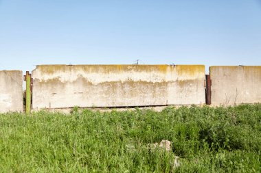 Section of a fence made of horizontal concrete slabs against a blue sky. Tall green grass in the foregroun