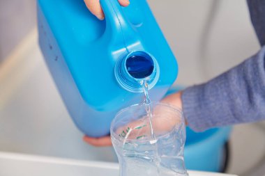 Hands pouring liquid from a blue plastic canister into a transparent container close u