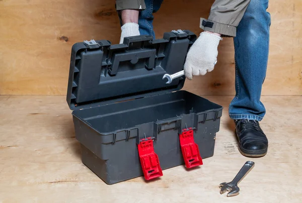 Gloved hands reach for tools from a modern plastic toolbox against a light-colored plywood backdrop. An adjustable wrench lies nearb