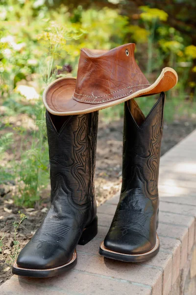 American farmer accessories. Black men cowboy-style leather boots stand on a brick path. Top brown leather ha