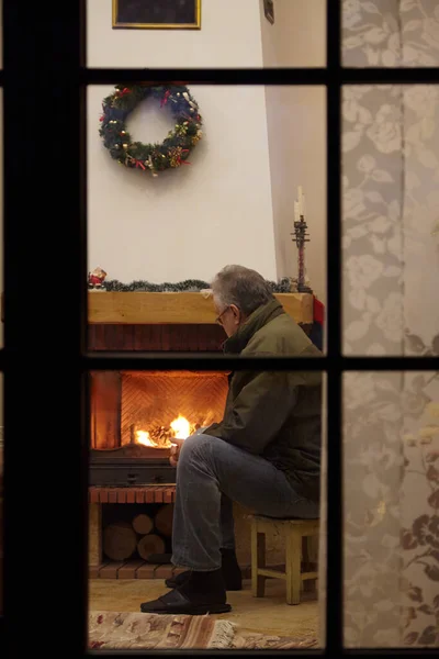 New Year Eve by fireplace. An elderly man sits on a carpet in front of fireplace in bright room. Fire burns brightly in the fireplace. Christmas wreath hangs over fireplace. View through windo