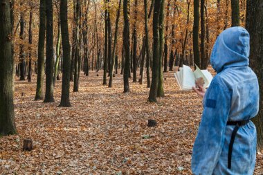 Gizemli sonbahar okumaları. Kapüşonlu bir kadın ağaçların arkasında kitap okuyor. Kahverengi yapraklar yerde yatıyor. Kitap ayracı yerine meşe tilkileri kullanılır.
