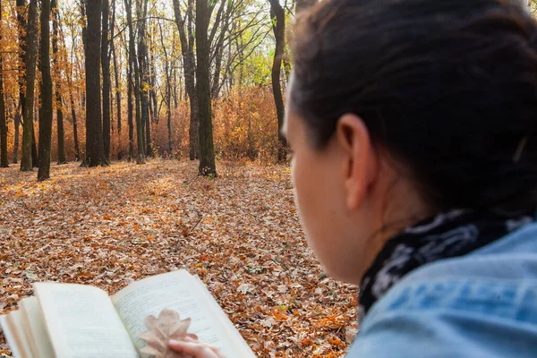 Romantic reading. A woman reads a book on a background of trees. Brown leaves are lying on the ground. Instead of a bookmark, oak foxes are used in the book