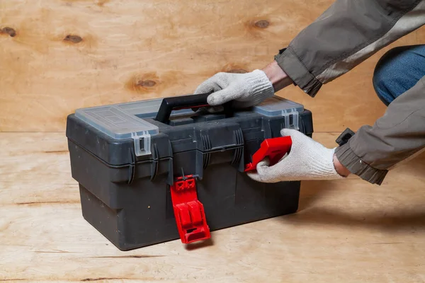 Hands in work gloves open a modern plastic closed toolbox on a light plywood backgroun