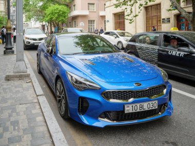 Mavi renkli Kia Stinger spor arabası Bakü caddesinde Uber taksiyle park etmiş.