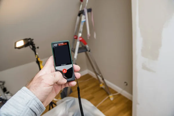 POV male hand holding Bosch laser meter in hand during renovation of a room ladder — стоковое фото