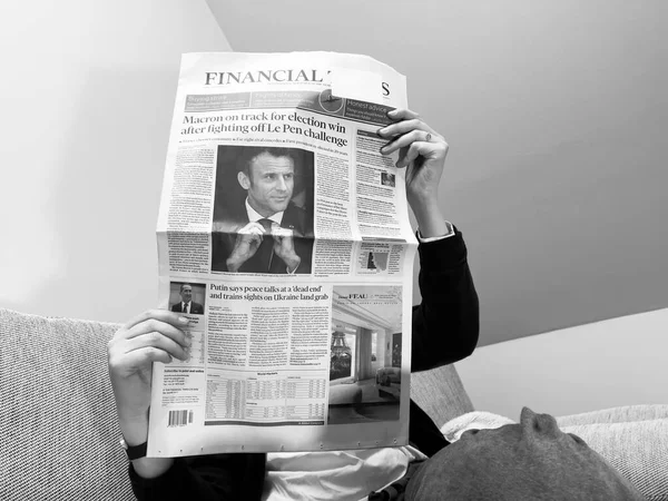 Mujer de imagen monocromática leyendo en un acogedor sofá el último periódico del Financial Times, cubrir con Frances presidente titular Emmanuel Macron después de vencer a Marine Le Pen por un segundo mandato de cinco años —  Fotos de Stock