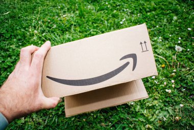 Yeşil çimlerin üzerinde gülümseyen ok logosu olan Amazon karton kutusunu tutan POV erkek.