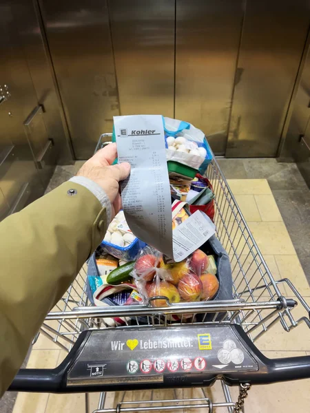 POV male hand holding supermarket receipt from Edeka Supermarket in the elevator of a shopping center looking at the rised prices of products in the cart — Foto de Stock