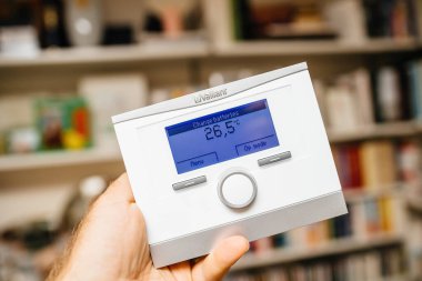 Remscheid, Germany - Jul 31, 2022: POV male hand holding new Vaillant VRC700 Weather compensating programmable room thermostat Intelligent control for heating and hot water - defocused room house