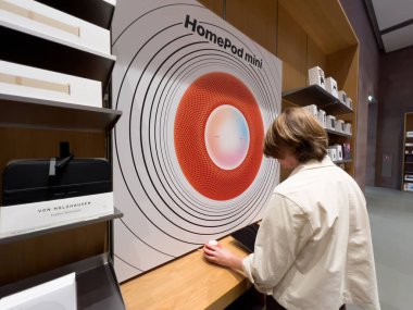 Paris, France - Sep 9, 2022: Woman looking at the new HomePod mini smart speaker in orange color with large poster inside Apple Store large advertising board for the product from Apple clipart