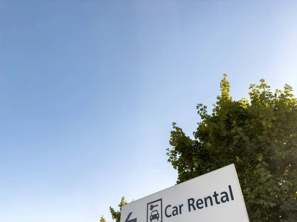 Low angle view of Car Rental sign with car and key pictogram as seen outside next to airport - tree with sunlight flare in background