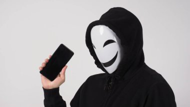 Anonim hacker ve yüz maskesi elinde akıllı telefonla. Siyah kapüşonlu tişörtlü adam beyaz arka planda cep telefonu kullanıyor. Siber suçlar veri hackleme veya kişisel veri çalma kavramını temsil et