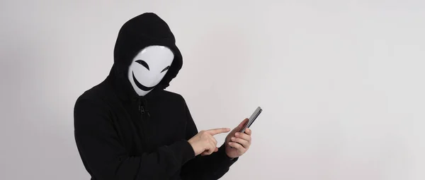 Anonymous hacker and face mask with smartphone in hand. Man in black hood shirt holding and using mobile phone on white background. Represent cyber crime data hacking or stealing personal data concept