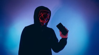 Anonim hacker ve yüz maskesi elinde akıllı telefonla. Siyah kapüşonlu bir adam cep telefonundan kişisel bilgileri hacklemeye çalışıyor. Siber suç veri hackleme veya kişisel veri çalma kavramını temsil et.