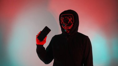 Anonim hacker ve yüz maskesi elinde akıllı telefonla. Siyah kapüşonlu bir adam cep telefonundan kişisel bilgileri hacklemeye çalışıyor. Siber suç veri hackleme veya kişisel veri çalma kavramını temsil et.