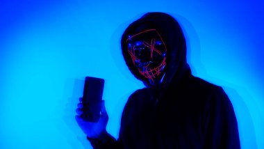 Anonim hacker ve yüz maskesi elinde akıllı telefonla. Siyah kapüşonlu bir adam cep telefonundan kişisel bilgileri hacklemeye çalışıyor. Siber suç veri hackleme veya kişisel veri çalma kavramını temsil et.