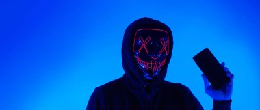 Anonim hacker ve yüz maskesi elinde akıllı telefonla. Siyah kapüşonlu bir adam cep telefonundan kişisel bilgileri hacklemeye çalışıyor. Siber suç veri hackleme veya kişisel veri çalma kavramını temsil et.