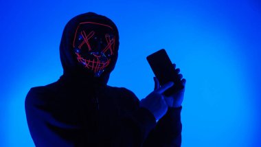 Anonim hacker ve yüz maskesi elinde akıllı telefonla. Siyah kapüşonlu bir adam cep telefonundan kişisel bilgileri hacklemeye çalışıyor. Siber suç veri hackleme veya kişisel veri çalma kavramını temsil et.