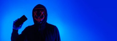 Anonim hacker ve yüz maskesi elinde akıllı telefonla. Siyah kapüşonlu bir adam cep telefonundan kişisel bilgileri hacklemeye çalışıyor. Siber suç veri hackleme veya kişisel veri çalma kavramını temsil et.