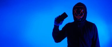 Anonim hacker ve yüz maskesi elinde akıllı telefonla. Siyah kapüşonlu bir adam cep telefonundan kişisel bilgileri hacklemeye çalışıyor. Siber suç veri hackleme veya kişisel veri çalma kavramını temsil et.