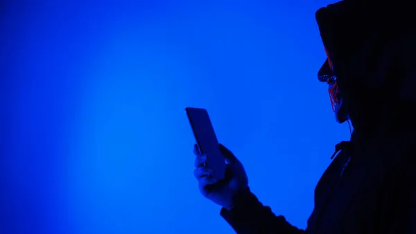 Anonymous hacker and face mask with smartphone in hand. Man in black hood shirt trying to hack personal data from mobile phone. Represent cyber crime data hacking or personal data stealing concept.