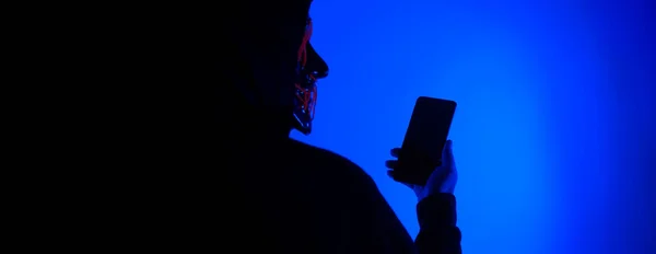 Anonymous hacker and face mask with smartphone in hand. Man in black hood shirt trying to hack personal data from mobile phone. Represent cyber crime data hacking or personal data stealing concept.