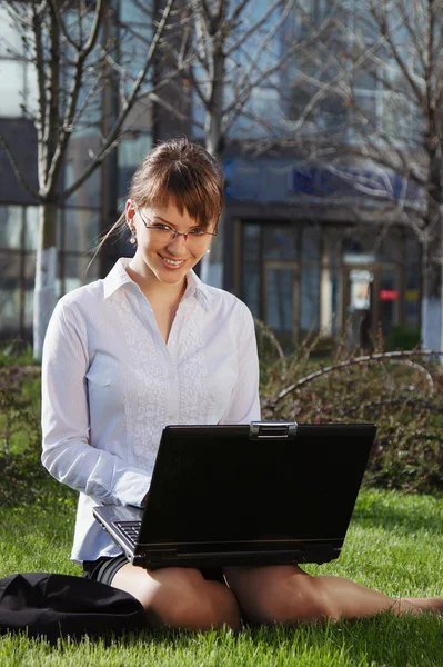 Woman lying on grass with laptop with business building on back — Stock Photo, Image