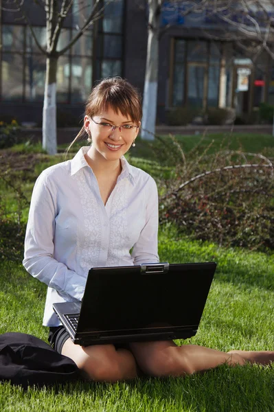Mujer acostada en la hierba con portátil con edificio de negocios en la espalda —  Fotos de Stock