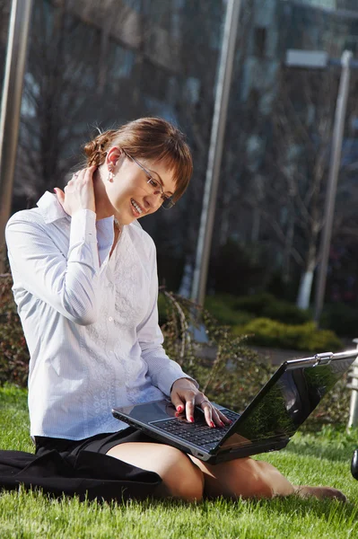 Жінка лежить на траві з ноутбуком з бізнес-будівлею на спині — стокове фото