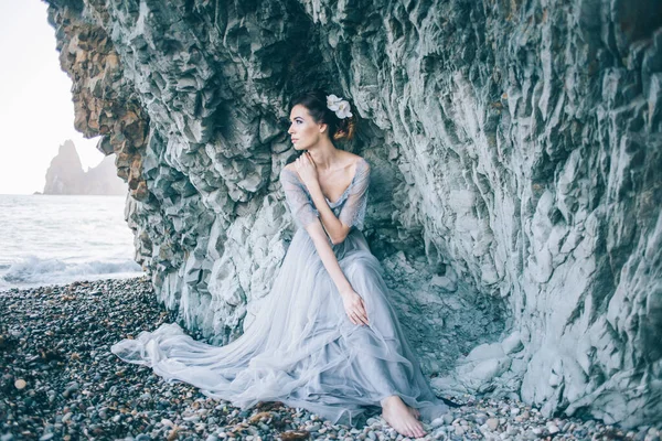美丽的黑发女孩在一个长灰色的礼服与面纱坐在海边 靠近蓝色的白色岩石查看剖面 — 图库照片