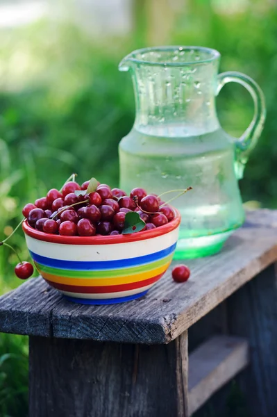 Jug of cold fresh water with cherries on wooden table in the garden — Stock Photo, Image