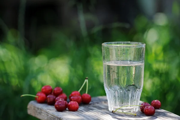 Glass of cold fresh water with cherries on wooden table in the garden — Stock Photo, Image