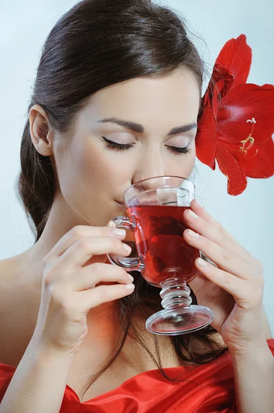 Hermosa joven con flor roja en el oído bebe té de frutas rojas de vidrio — Foto de Stock