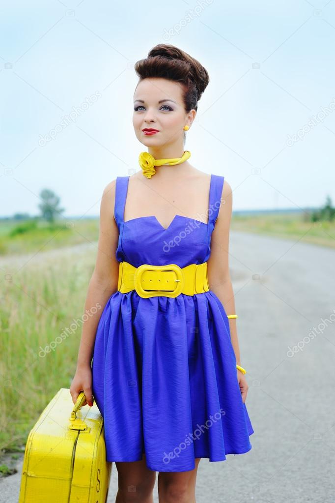 Beautiful smiling girl in a blue dress with a yellow suitcase on the road